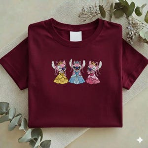 Puede incluir: Camiseta burdeos con un dise&ntilde;o bordado de tres personajes de dibujos animados con vestidos de princesa. Los personajes son rosas y azules con coronas. La camiseta tiene cuello redondo y mangas cortas.