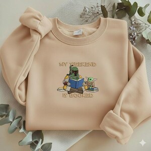 Pu&ograve; includere: Felpa beige con la frase ricamata "MY WEEKEND IS BOOKED" e una grafica di un personaggio che legge un libro con una piccola creatura verde. La felpa ha girocollo e maniche lunghe.