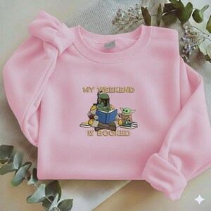 Pu&ograve; includere: Una felpa rosa chiaro con la scritta dorata "MY WEEKEND IS BOOKED". La grafica presenta un cartone animato di Boba Fett che legge un libro con Grogu. La felpa ha maniche lunghe e girocollo.
