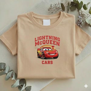 Puede incluir: Una camiseta beige con un dise&ntilde;o de Rayo McQueen de la pel&iacute;cula Cars. El dise&ntilde;o incluye el texto "LIGHTNING McQUEEN" en rojo, con la imagen del coche debajo. La camiseta est&aacute; doblada y colocada sobre una superficie neutra.