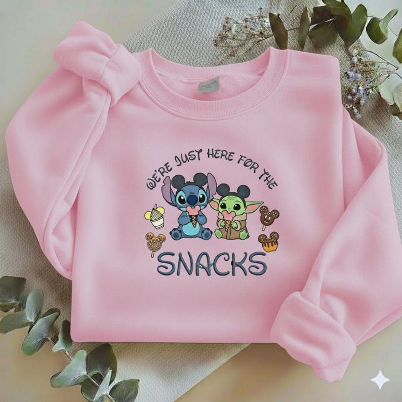 May include: A light pink sweatshirt featuring the characters Stitch and Grogu, with the text "WE'RE JUST HERE FOR THE SNACKS". The design includes cartoon snacks and the characters holding ice cream cones. The sweatshirt has long sleeves.