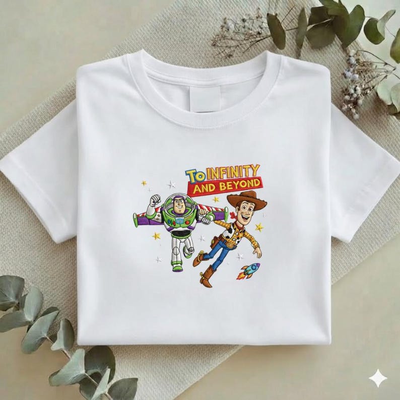 K&ouml;nnte beinhalten: Wei&szlig;es T-Shirt mit einer farbenfrohen Grafik von Buzz Lightyear und Woody aus Toy Story, mit dem Schriftzug "To Infinity and Beyond". Das Design beinhaltet eine Rakete und Sternakzente.