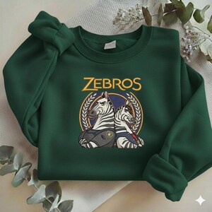 Może przedstawiać: Ciemnozielona bluza z napisem "ZEBROS" złotymi literami nad grafiką dw&oacute;ch zebr w okrągłym wzorze. Zebry mają na sobie kamizelki i poważny wyraz twarzy.