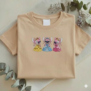 Puede incluir: Una camiseta beige con un dise&ntilde;o bordado de tres personajes de dibujos animados con vestidos de princesa. Los personajes son rosas con coronas y visten vestidos amarillos, azules y rosas. La camiseta est&aacute; doblada sobre una superficie beige claro.