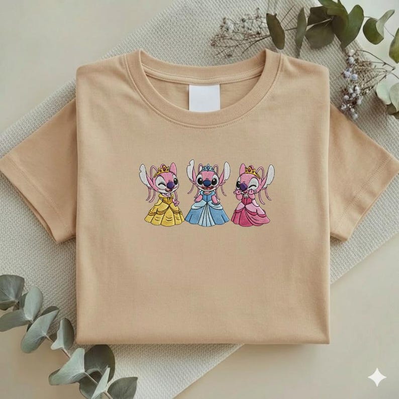 Puede incluir: Una camiseta beige con un dise&ntilde;o bordado de tres personajes de dibujos animados con vestidos de princesa. Los personajes son rosas con coronas y visten vestidos amarillos, azules y rosas. La camiseta est&aacute; doblada sobre una superficie beige claro.