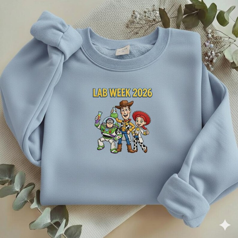 May include: A light blue sweatshirt featuring a graphic of three cartoon characters and the text "LAB WEEK 2026" in yellow. The graphic includes a space ranger, a cowboy, and a cowgirl. The sweatshirt has a crew neck and long sleeves.
