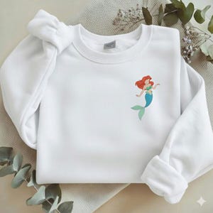 May include: White sweatshirt featuring a colorful illustration of a mermaid with red hair, a green top, and a blue tail. The sweatshirt has a crew neck and long sleeves with ribbed cuffs.