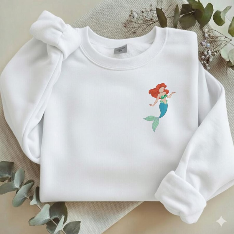 May include: White sweatshirt featuring a colorful illustration of a mermaid with red hair, a green top, and a blue tail. The sweatshirt has a crew neck and long sleeves with ribbed cuffs.