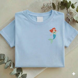 May include: A light blue t-shirt featuring a cartoon mermaid with red hair, a green top, and a blue tail. The shirt is folded neatly on a beige surface, with sprigs of greenery in the background.