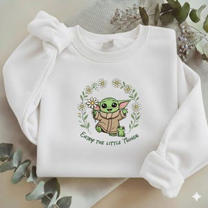 May include: White sweatshirt featuring a green cartoon character holding daisies, with a frog and the text "Enjoy the little things." The design is surrounded by a floral wreath. The sweatshirt has long sleeves.