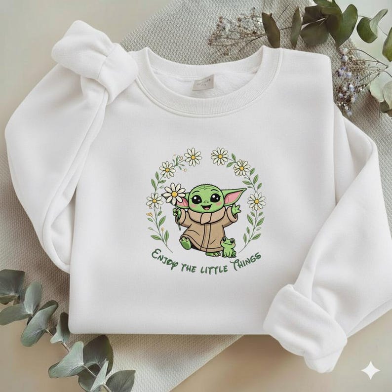 May include: White sweatshirt featuring a green cartoon character holding daisies, with a frog and the text "Enjoy the little things." The design is surrounded by a floral wreath. The sweatshirt has long sleeves.