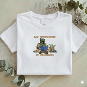 Pu&ograve; includere: T-shirt bianca con una grafica di un personaggio con un'armatura verde che legge un libro blu, con una piccola creatura verde. Il testo "MY WEEKEND IS BOOKED" &egrave; sopra e sotto i personaggi.