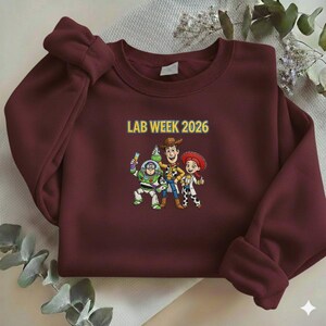 May include: A maroon sweatshirt featuring a graphic of three cartoon characters and the text "LAB WEEK 2026" in yellow. The cartoon characters are Buzz Lightyear, Woody, and Jessie. The sweatshirt is made of a soft material.