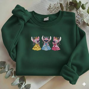 Puede incluir: Sudadera verde oscuro con un dise&ntilde;o bordado de tres personajes de dibujos animados con vestidos de princesa. Los personajes son rosas con coronas y visten vestidos amarillos, azules y rosas. La sudadera tiene mangas largas y cuello redondo.