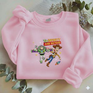 K&ouml;nnte beinhalten: Hellrosa Sweatshirt mit einem Aufdruck von Buzz Lightyear und Woody aus Toy Story, mit dem Text "TO INFINITY AND BEYOND". Das Design beinhaltet Sterne und eine kleine Rakete. Das Sweatshirt hat lange &Auml;rmel.