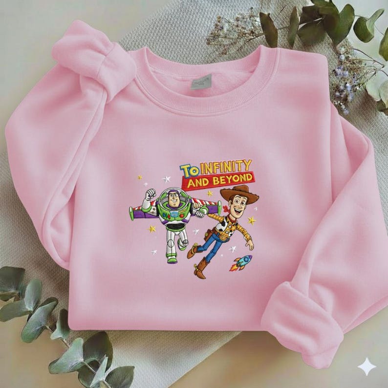 K&ouml;nnte beinhalten: Hellrosa Sweatshirt mit einem Aufdruck von Buzz Lightyear und Woody aus Toy Story, mit dem Text "TO INFINITY AND BEYOND". Das Design beinhaltet Sterne und eine kleine Rakete. Das Sweatshirt hat lange &Auml;rmel.
