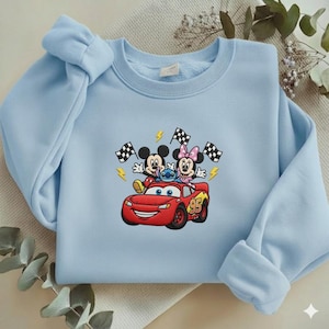 May include: Light blue sweatshirt featuring an embroidered design of Mickey Mouse, Minnie Mouse, and Stitch riding in a red race car. The design includes checkered flags and lightning bolts. The sweatshirt has a crew neck and long sleeves.