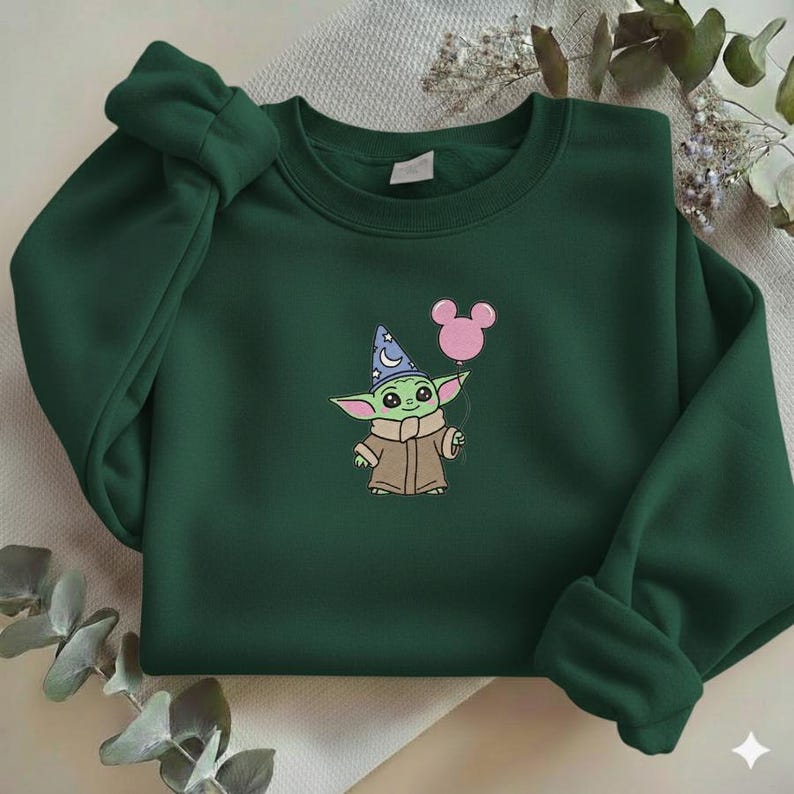 Op de afbeelding: Donkergroene sweatshirt met een geborduurd stripfiguur met een tovenaars hoed en een roze ballon. Het personage is lichtgroen met grote ogen en een bruine jas. De sweatshirt heeft lange mouwen en een ronde hals.