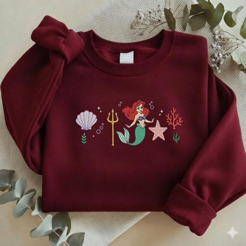 Puede incluir: Sudadera burdeos con un dise&ntilde;o bordado de una sirena con pelo rojo y cola verde, un tridente dorado, una concha y una estrella de mar. La sudadera tiene mangas largas y cuello redondo.