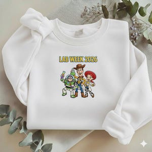 May include: White crewneck sweatshirt featuring a colorful graphic of three cartoon characters and the text "LAB WEEK 2026" in yellow. The graphic includes a space ranger, a cowboy, and a cowgirl.