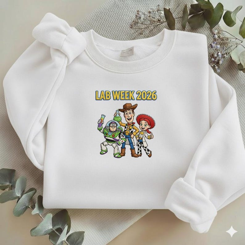 May include: White crewneck sweatshirt featuring a colorful graphic of three cartoon characters and the text "LAB WEEK 2026" in yellow. The graphic includes a space ranger, a cowboy, and a cowgirl.