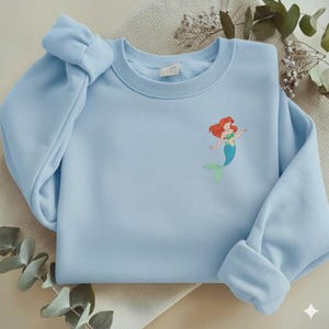 May include: A light blue sweatshirt featuring an embroidered image of a mermaid with red hair, a green top, and a blue tail. The sweatshirt has long sleeves and a crew neck. The sleeves are cuffed at the wrists.