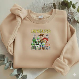 Puede incluir: Una sudadera beige con el texto "LAB WEEK 2026" sobre un gr&aacute;fico de personajes de dibujos animados. El gr&aacute;fico incluye un vaquero, un guardabosques espacial, una vaquera, un cerdo y otros personajes con equipo de laboratorio.