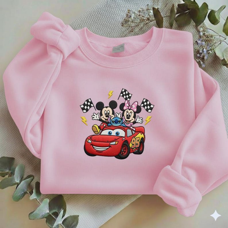 May include: A light pink sweatshirt featuring a colorful graphic of Mickey Mouse, Minnie Mouse, and Stitch riding in a red race car. The design includes checkered flags and lightning bolts. The car has the number 95 on the side.