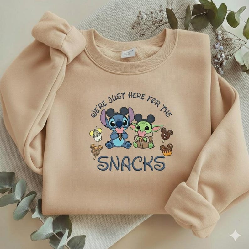 May include: A beige sweatshirt featuring an embroidered design of Stitch and Grogu with the text "We're just here for the snacks." The design includes images of snacks and ice cream cones. The sweatshirt has a crew neck and long sleeves.