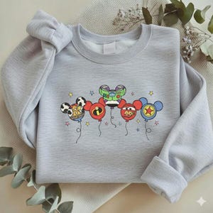 Może przedstawiać: Jasnoszara bluza z kolorową grafiką balon&oacute;w z postaciami. Balony zawierają wzory Myszki Miki, Buzz Lightyear i Zygzaka McQueena. Bluza ma długie rękawy i okrągły dekolt.