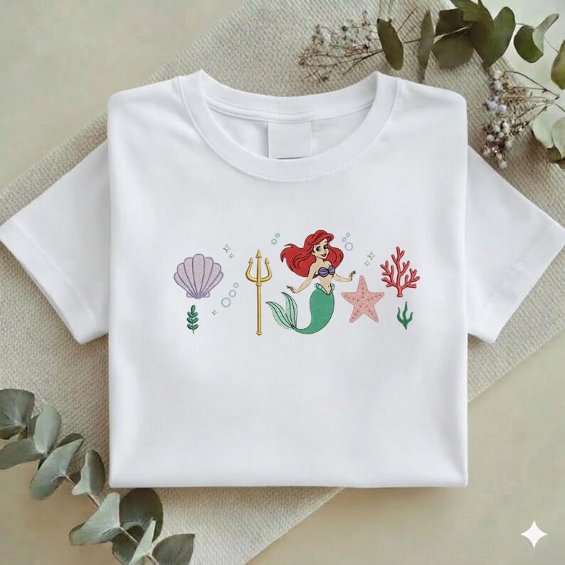 Puede incluir: Camiseta blanca con un dise&ntilde;o bordado. El dise&ntilde;o presenta una sirena con cabello rojo y cola verde, un tridente dorado, una estrella de mar rosa y una concha. Tambi&eacute;n se incluyen corales y burbujas.
