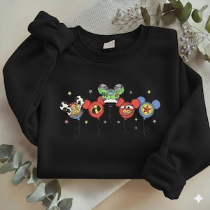 Może przedstawiać: Czarna bluza z haftowanymi balonami z postaciami z kresk&oacute;wek. Balony zaprojektowano jako głowy Myszki Miki, z kt&oacute;rych każda reprezentuje inną postać. Postacie obejmują krowę, Buzz Lightyear i Zygzaka McQueena.