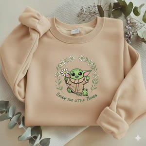 May include: Beige sweatshirt featuring a green cartoon character holding a daisy, surrounded by a floral wreath. The text "Enjoy the little things" is printed below the character. The sweatshirt has a crew neck and long sleeves.
