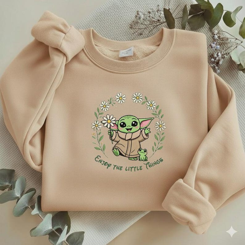 May include: Beige sweatshirt featuring a green cartoon character holding a daisy, surrounded by a floral wreath. The text "Enjoy the little things" is printed below the character. The sweatshirt has a crew neck and long sleeves.