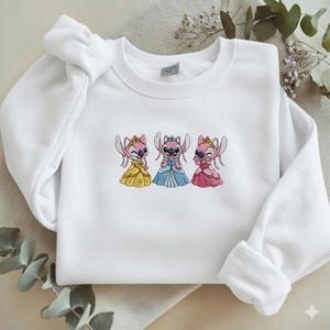 Puede incluir: Sudadera blanca con bordados de personajes Stitch con vestidos de princesa. El dise&ntilde;o incluye a Stitch con vestidos amarillos, azules y rosas, cada uno con una corona. La sudadera tiene cuello redondo y mangas largas.
