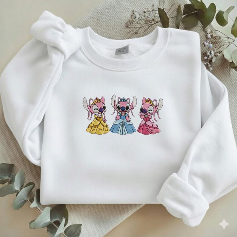 Puede incluir: Sudadera blanca con bordados de personajes Stitch con vestidos de princesa. El dise&ntilde;o incluye a Stitch con vestidos amarillos, azules y rosas, cada uno con una corona. La sudadera tiene cuello redondo y mangas largas.