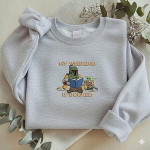 Pu&ograve; includere: Felpa grigio chiaro con la frase ricamata "MY WEEKEND IS BOOKED". Il design presenta un personaggio dei cartoni animati con un'armatura verde che legge un libro blu con una piccola creatura verde. La felpa ha maniche lunghe.