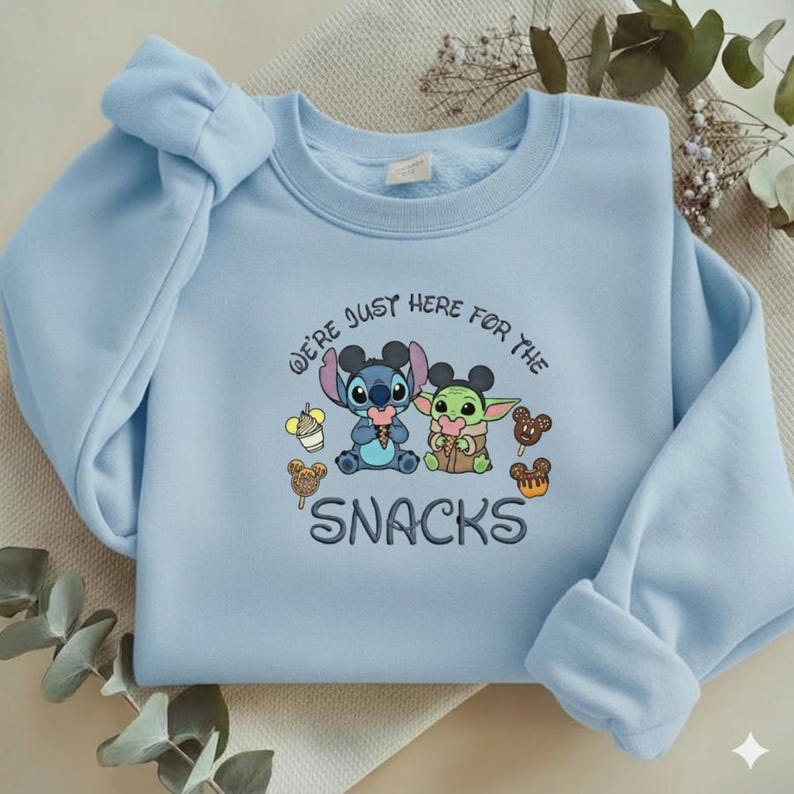 May include: Light blue sweatshirt featuring embroidered cartoon characters Stitch and Grogu, with the text "WE'RE JUST HERE FOR THE SNACKS". The design includes ice cream and Mickey Mouse-shaped treats.