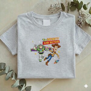K&ouml;nnte beinhalten: Hellgraues T-Shirt mit einem Aufdruck von Buzz Lightyear und Woody aus Toy Story. Die Grafik enth&auml;lt den Schriftzug "To Infinity and Beyond" in Gelb und Rot. Die Charaktere sind in Bewegung dargestellt, mit einer kleinen Rakete.