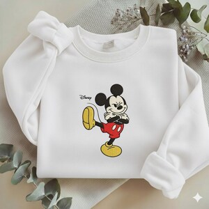 Puede incluir: Sudadera blanca de cuello redondo con un dise&ntilde;o bordado de Mickey Mouse. Mickey se muestra con los brazos cruzados y una pierna levantada. El logotipo de Disney est&aacute; impreso en el lado izquierdo de la sudadera.