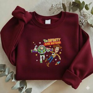 K&ouml;nnte beinhalten: Ein weinrotes Sweatshirt mit einer farbenfrohen Grafik von Buzz Lightyear und Woody aus Toy Story, mit dem Text "To Infinity and Beyond". Das Design umfasst Sterne und eine Rakete.