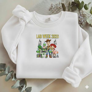 Puede incluir: Sudadera blanca con un gr&aacute;fico colorido que presenta personajes de la pel&iacute;cula Toy Story, incluyendo a Woody, Buzz Lightyear, y otros, con el texto "LAB WEEK 2026" en amarillo.