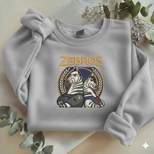 Może przedstawiać: Jasnoszara bluza z okrągłym dekoltem i długimi rękawami. Zawiera kresk&oacute;wkowy wz&oacute;r dw&oacute;ch zebr w policyjnych mundurach, ze złotym napisem "ZEBROS" powyżej. Zebry umieszczone są w niebiesko-złotym emblemacie.