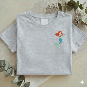 May include: A light gray t-shirt featuring a small embroidered image of a mermaid with red hair, a green top, and a blue tail. The shirt is folded neatly on a neutral-colored surface.