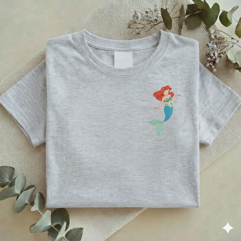 May include: A light gray t-shirt featuring a small embroidered image of a mermaid with red hair, a green top, and a blue tail. The shirt is folded neatly on a neutral-colored surface.
