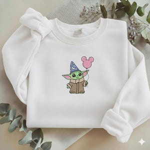 Op de afbeelding: Witte sweatshirt met een geborduurd stripfiguur met een tovenaars hoed en een roze ballon. Het karakter is groen met grote ogen en een bruine jas. De sweatshirt heeft lange mouwen en een ronde hals.