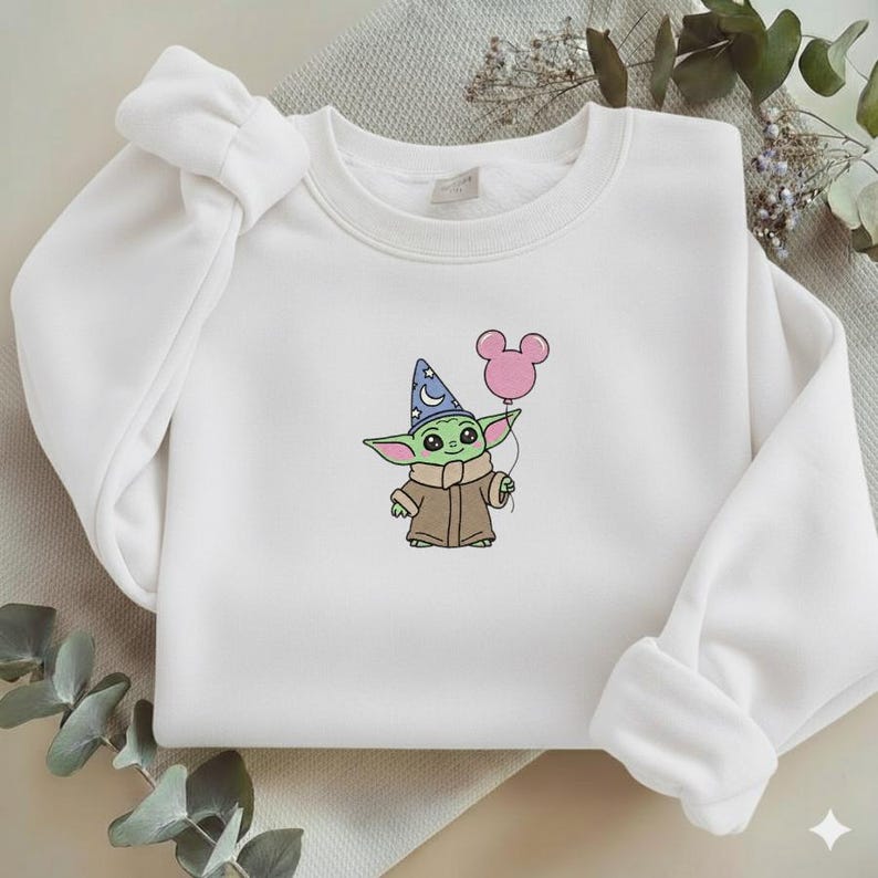 Op de afbeelding: Witte sweatshirt met een geborduurd stripfiguur met een tovenaars hoed en een roze ballon. Het karakter is groen met grote ogen en een bruine jas. De sweatshirt heeft lange mouwen en een ronde hals.