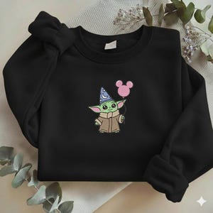 Op de afbeelding: Zwart sweatshirt met een geborduurd beeld van een groen alien-personage met een tovenaars hoed en een roze ballon. Het personage draagt een bruine jas. De ballon heeft de vorm van Mickey Mouse oren.