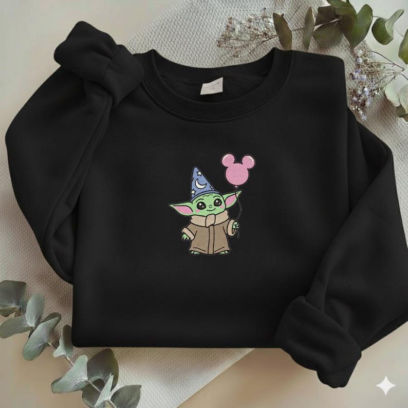 Op de afbeelding: Zwart sweatshirt met een geborduurd beeld van een groen alien-personage met een tovenaars hoed en een roze ballon. Het personage draagt een bruine jas. De ballon heeft de vorm van Mickey Mouse oren.