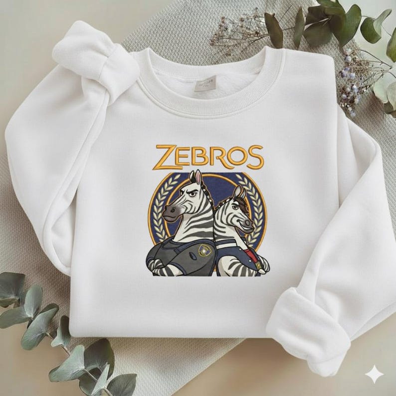 Może przedstawiać: Biała bluza z nadrukiem dw&oacute;ch zebr w okrągłym emblemacie z napisem "ZEBROS" powyżej. Zebry są w stylu kresk&oacute;wkowym, jedna z nich ma na sobie mundur. Projekt wykorzystuje kolory niebieski, złoty i czarny.